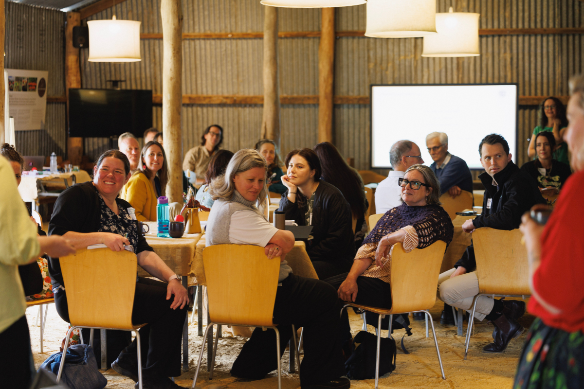Group of people seated at round tables in a rustic indoor setting, attentively listening to a speaker off camera.